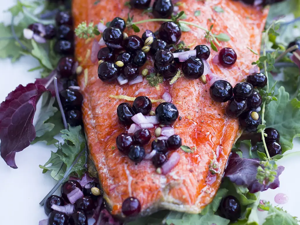 Cooked salmon topped with fresh huckleberries, red onions, and herbs on a bed of leafy greens.