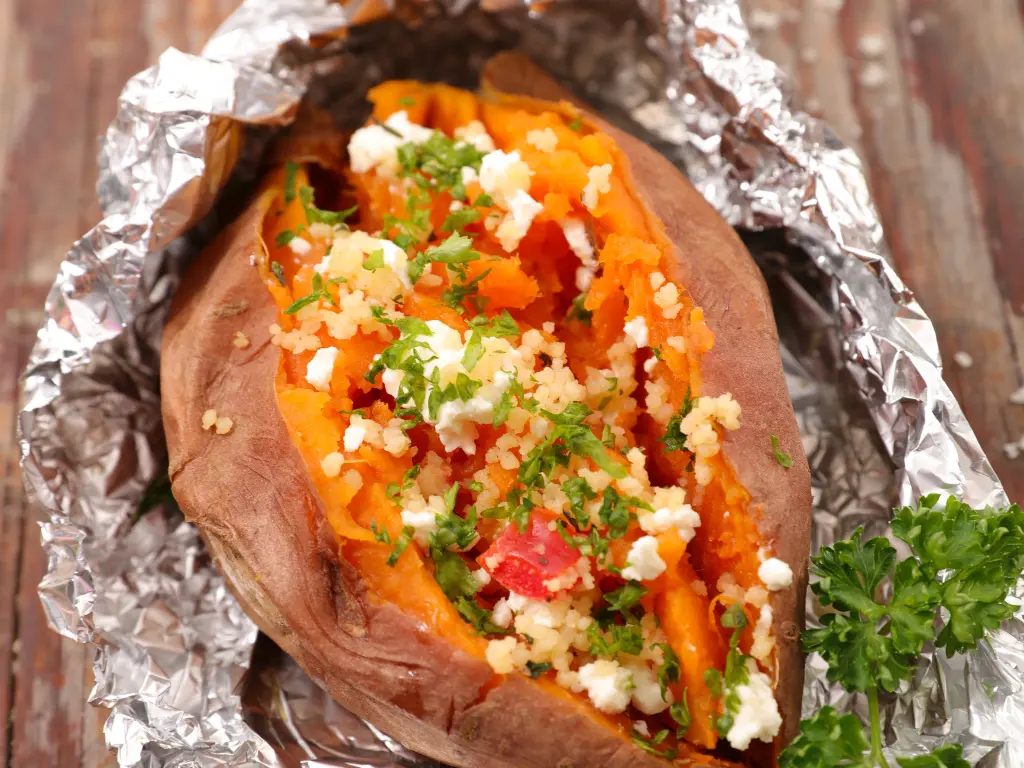 Cooked Beauregard sweet potato split open and filled with couscous, cheese, and parsley on foil wrap.